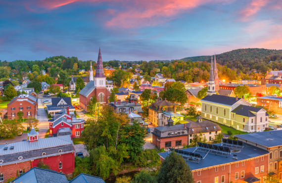 Aerial view of a small town at dusk, with two illuminated church steeples, red brick buildings, and green hillsides in the background.